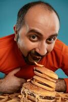 Close-up portrait of a middle-aged man with beard, dressed in a red turtleneck, posing with burgers and french fries. Blue background. Fast food. photo