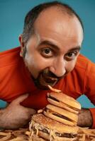 de cerca retrato de un de edad mediana hombre con barba, vestido en un rojo suéter tipo con cuello de tortuga, posando con hamburguesas y francés papas fritas azul antecedentes. rápido alimento. foto