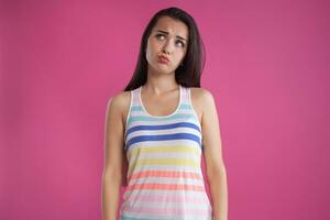 Brunette woman with long hair, dressed in colorful striped shirt, posing against pink studio background. Sincere emotions. Close-up. photo