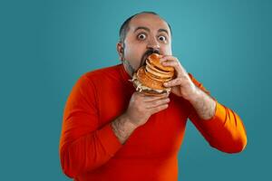 Close-up portrait of a middle-aged man with beard, dressed in a red turtleneck, posing with burgers against a blue background. Fast food. photo