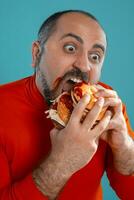 de cerca retrato de un de edad mediana hombre con barba, vestido en un rojo suéter tipo con cuello de tortuga, posando con hamburguesas en contra un azul antecedentes. rápido alimento. foto