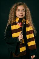 Portrait of a little witch girl with long brown hair dressed in dark coat, holding magic wand in her hand, posing on black studio background. photo