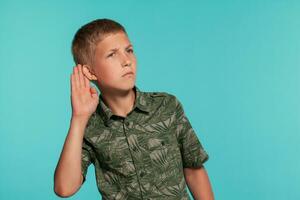 Close-up portrait of a blonde teenage boy in a green shirt with palm print posing against a blue studio background. Concept of sincere emotions. photo