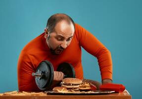 Close-up portrait of a middle-aged man with beard, dressed in a red turtleneck, posing with burgers and french fries. Blue background. Fast food. photo