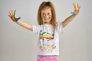 Little girl in white painted t-shirt is posing standing isolated on white and gesticulating with her colored in different paints palms and face. Art studio. Close-up. photo