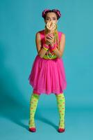 Lovely girl with a multi-colored braids hairstyle and bright make-up, posing in studio against a blue background, holding a lollipop in her hand. photo