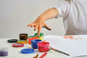 Little artist in white t-shirt standing at table with whatman and colorful paints, painting on it with her hands. Isolated on white. Close-up. photo