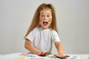 Little girl in white t-shirt sitting at table with whatman and paints on it, posing with painted face and hands. Isolated on white. Medium close-up. photo