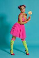 Lovely girl with a multi-colored braids hairstyle and bright make-up, posing in studio against a blue background, holding a lollipop in her hand. photo