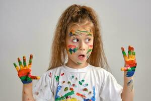 Little girl in white t-shirt is posing standing isolated on white and gesticulating with her painted in different colors palms. Art studio. Close-up. photo