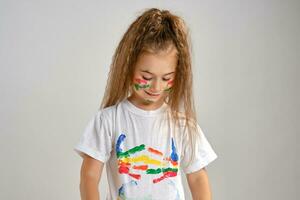 Little girl in white painted t-shirt is posing standing isolated on white, looking down. Her face colored in different paints. Art studio. Close-up. photo