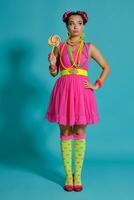 Lovely girl with a multi-colored braids hairstyle and bright make-up, posing in studio against a blue background, holding a lollipop in her hand. photo