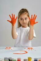 Little girl in white t-shirt sitting at table with whatman and colorful paints, showing her painted hands. Isolated on white. Medium close-up. photo