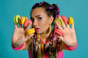 Lovely girl with multi-colored braids hairstyle and bright make-up, holding macarons between her fingers, posing in studio against a blue background. photo