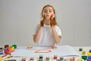 pequeño niña en blanco camiseta sentado a mesa con qué hombre y vistoso pinturas en él, pintura su las mejillas. aislado en blanco. medio de cerca. foto