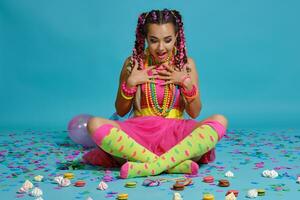 Lovely girl with a multi-colored braids hairstyle and bright make-up, posing in studio with air balloons and confetti against a blue background. photo