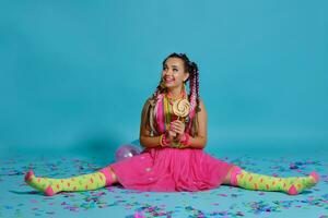 Lovely girl with a multi-colored braids hairstyle and bright make-up, posing in studio with lollipop, air balloons and confetti against a blue background. photo