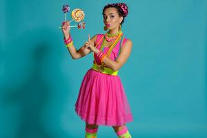 encantador niña con un multicolor trenzas peinado y brillante constituir, posando en estudio en contra un azul fondo, participación un pirulí en su mano. foto