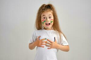 Little girl in white painted t-shirt is posing standing isolated on white and gesticulating with her colored in different paints palms and face. Art studio. Close-up. photo