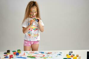 Little girl in white t-shirt standing at table with whatman and colorful paints,playing with a sponge soaked in paint. Isolated on white. Close-up. photo