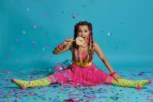 Lovely girl with a multi-colored braids hairstyle and bright make-up, posing in studio with lollipop, air balloons and confetti against a blue background. photo