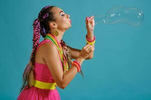 encantador niña con un multicolor trenzas peinado y brillante constituir, es soplo burbujas utilizando su manos, posando en estudio en contra un azul antecedentes. foto