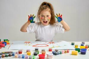 Little girl in white t-shirt sitting at table with whatman and paints on it, posing with painted face and hands. Isolated on white. Medium close-up. photo