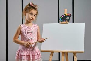 pequeño niña artista en un rosado vestir es en pie detrás caballete y pintura con cepillo en lona a Arte estudio con blanco paredes medio de cerca disparo. foto