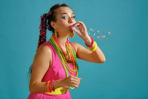 encantador niña con un multicolor trenzas peinado y brillante constituir, es soplo burbujas utilizando su manos, posando en estudio en contra un azul antecedentes. foto