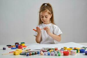 Little girl in white t-shirt sitting at table with whatman and colorful paints on it, painting her hands. Isolated on white. Medium close-up. photo