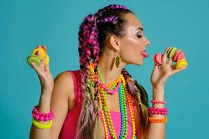 encantador niña con multicolor trenzas peinado y brillante constituir, participación macarons Entre su dedos, posando en estudio en contra un azul antecedentes. foto