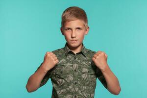 Close-up portrait of a blonde teenage boy in a green shirt with palm print posing against a blue studio background. Concept of sincere emotions. photo