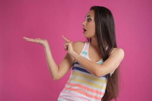 Brunette woman with long hair, dressed in colorful striped shirt, posing against pink studio background. Sincere emotions. Close-up. photo