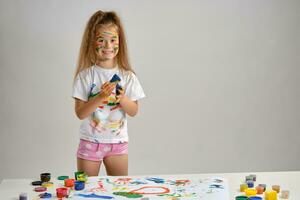 Little girl in white t-shirt standing at table with whatman and colorful paints, playing with a sponge soaked in paint. Isolated on white. Close-up. photo