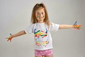 Little girl in white painted t-shirt is posing standing isolated on white and gesticulating with her colored in different paints palms and face. Art studio. Close-up. photo