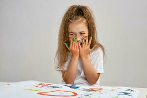 Little girl in white t-shirt sitting at table with whatman and paints on it, posing with painted face and hands. Isolated on white. Medium close-up. photo