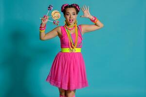 Lovely girl with a multi-colored braids hairstyle and bright make-up, posing in studio against a blue background, holding a lollipop in her hand. photo