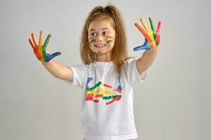 Little girl in white painted t-shirt is posing standing isolated on white and gesticulating with her colored in different paints palms and face. Art studio. Close-up. photo