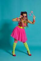 encantador niña con un multicolor trenzas peinado y brillante constituir, posando en estudio en contra un azul fondo, participación un pirulí en su mano. foto