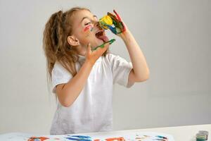 Little girl in white t-shirt sitting at table with whatman and paints on it, posing with painted face and hands. Isolated on white. Medium close-up. photo