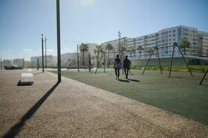 un sencillo al aire libre urbano campo deportivo en el paseo en contra el antecedentes de moderno edificios antecedentes. posterior ver de irreconocible mujer caminando abajo el calle en soleado día foto