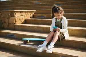 Adorable child girl using mobile phone, browses internet, plays online video games, watches cartoons, sitting on steps photo