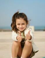 Authentic charming little child girl showing thumbs up and smiling looking at camera, sitting on her skateboard outdoors photo