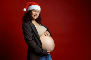 encantador embarazada mujer en rojo Papa Noel sombrero, suavemente acariciando su grande barriga, sonriente mirando a cámara aislado rojo fondo foto