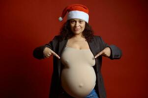 embarazada mujer en Papa Noel sombrero, señalando dedos a su grande barriga en el embarazo tercero trimestre, aislado en rojo antecedentes. foto
