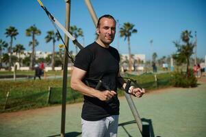 Confident portrait of handsome Caucasian sportsman smiling looking at camera, exercising with suspension straps outdoors photo