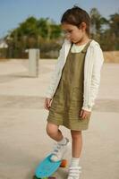 Little kid girl standing on skateboard on one leg, expressing sad emotions having difficulties on learning skateboarding photo