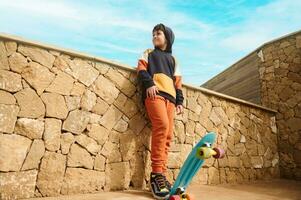 Full length portrait of handsome teenager boy in sportswear, playing with a skateboard, looking aside photo