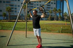 Young adult sportsman doing bodyweight exercises on arms, push ups with suspension straps in urban sports ground photo