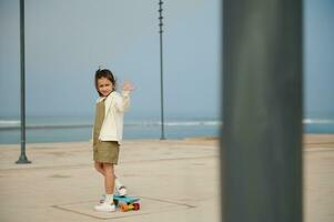 adorable pequeño niño niña saludo con mano, en pie en su azul patineta en uno pierna en un moderno urbano patinar parque foto